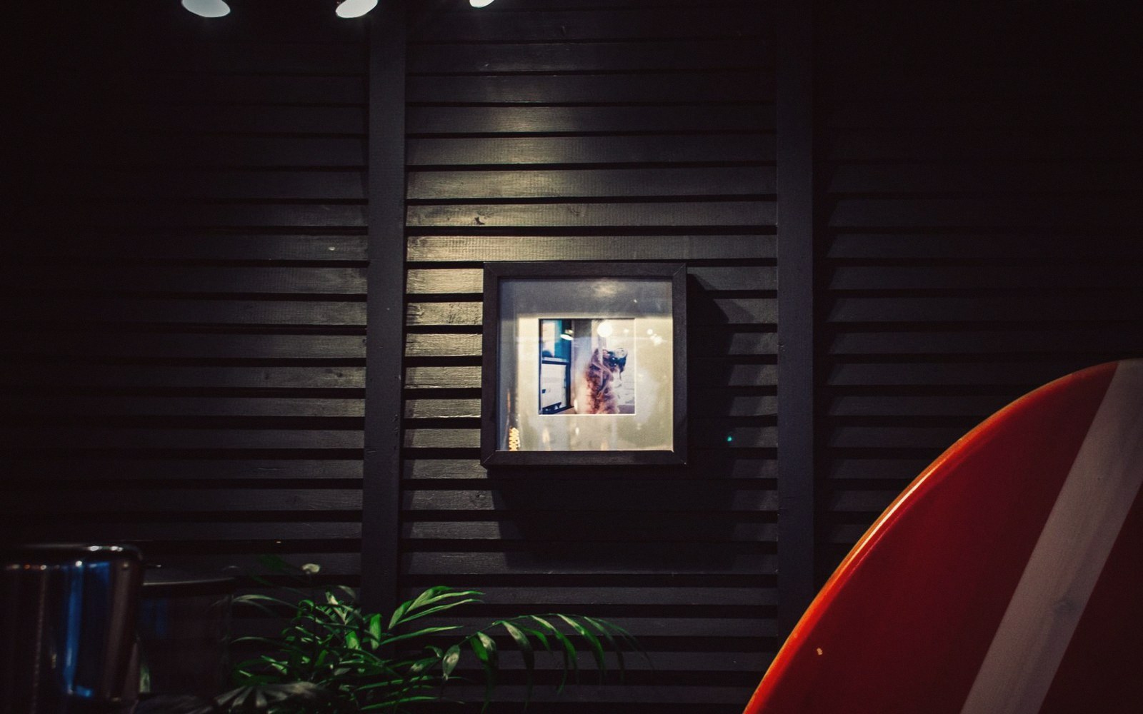 Framed photo glowing on a dark slatted wall beside a red surfboard and plant