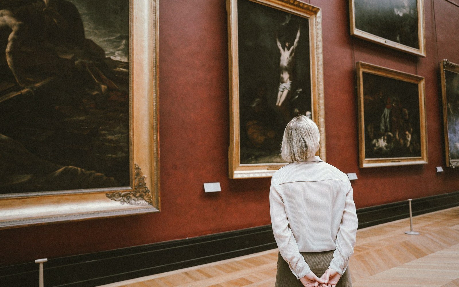 A woman views framed paintings on a red gallery wall from behind