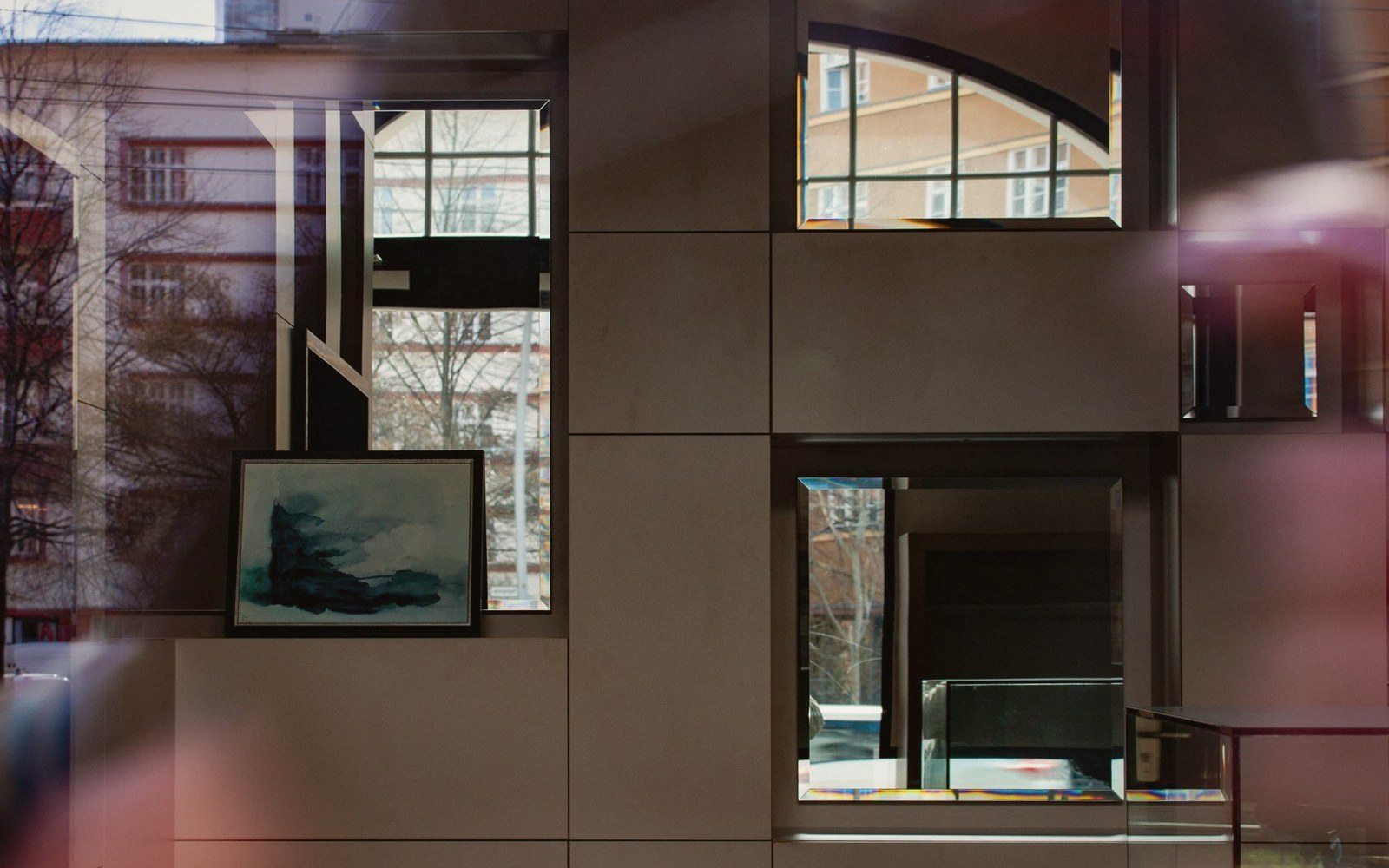 A framed painting sits on a shelf inside a glass-walled room reflecting nearby buildings