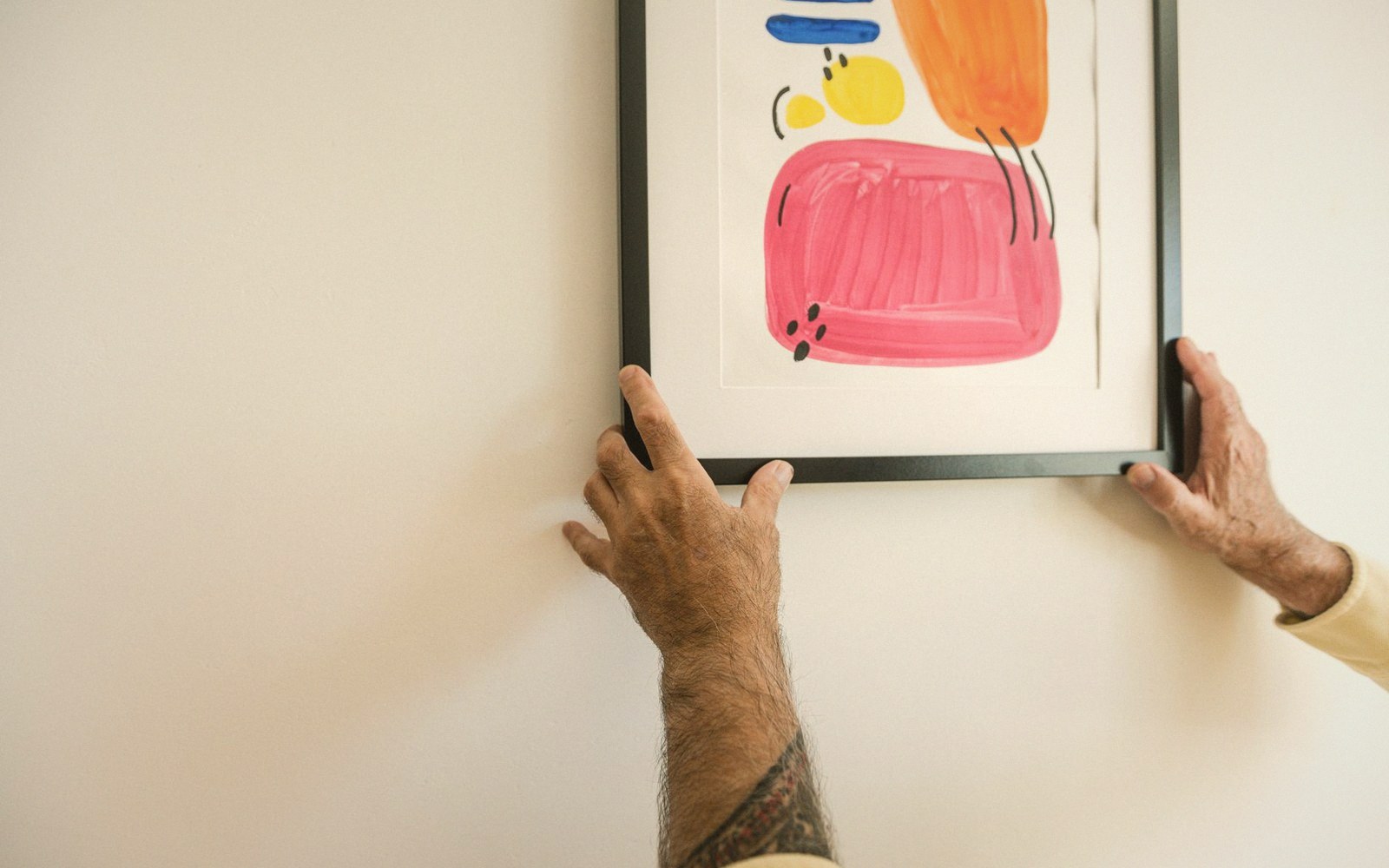 Hands positioning a framed abstract painting with pink, orange, yellow and blue shapes on a wall