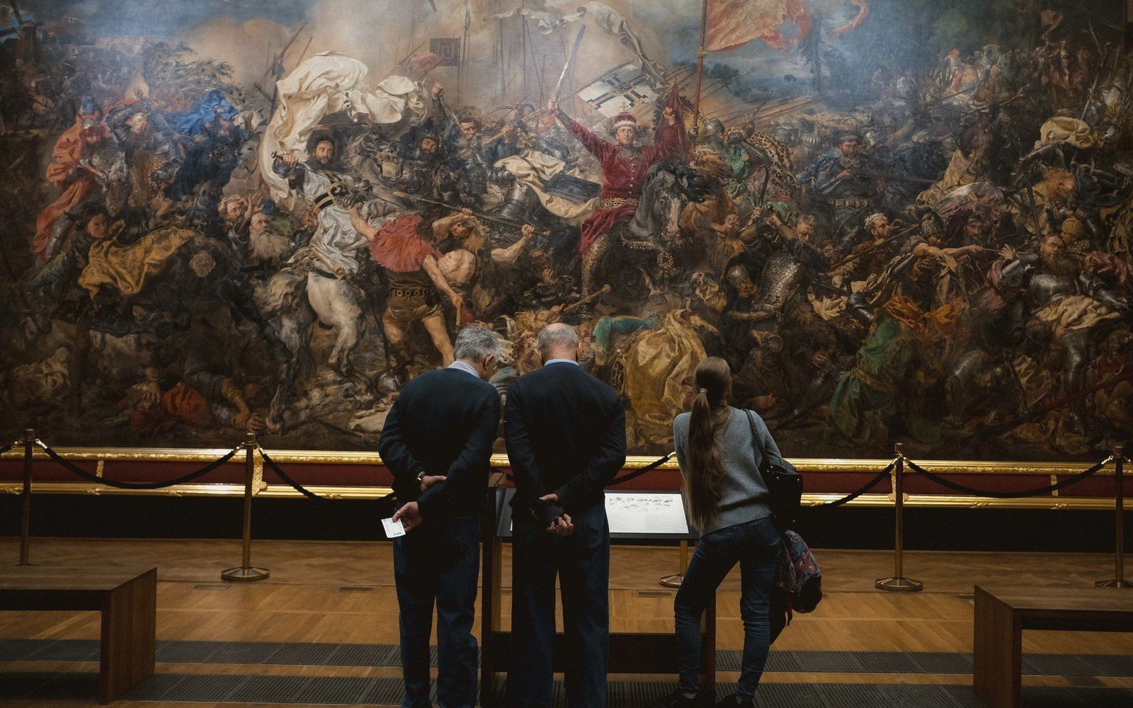 Visitors study a large historical battle painting depicting mounted knights and soldiers in combat