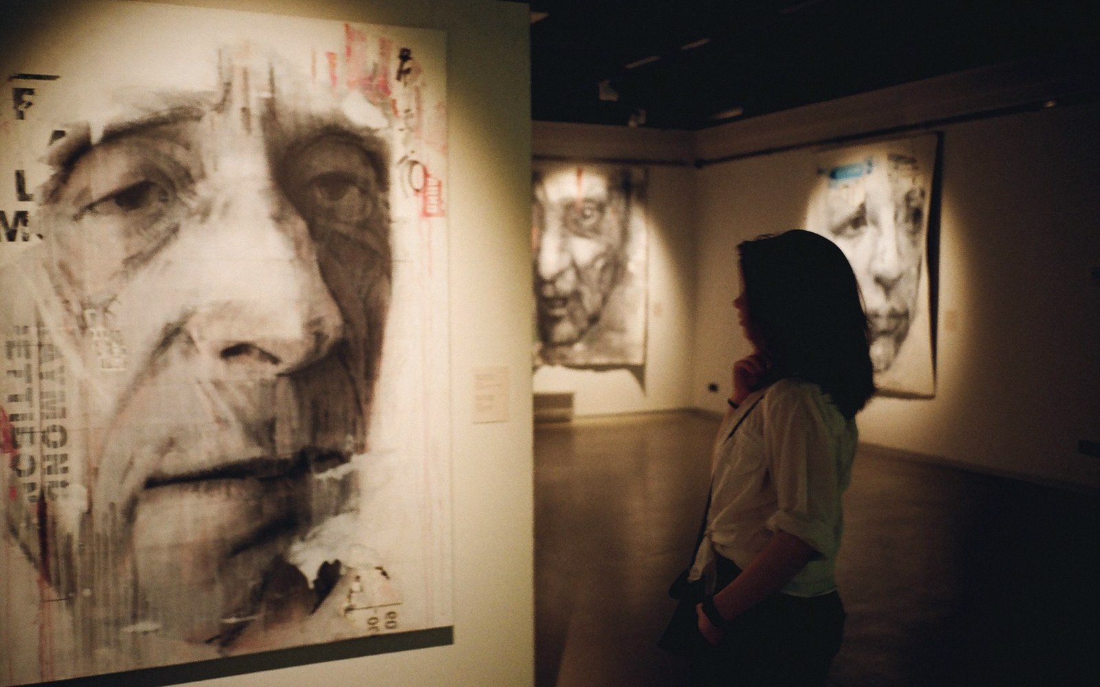 A visitor studies large charcoal portrait paintings hung in a dimly lit gallery room